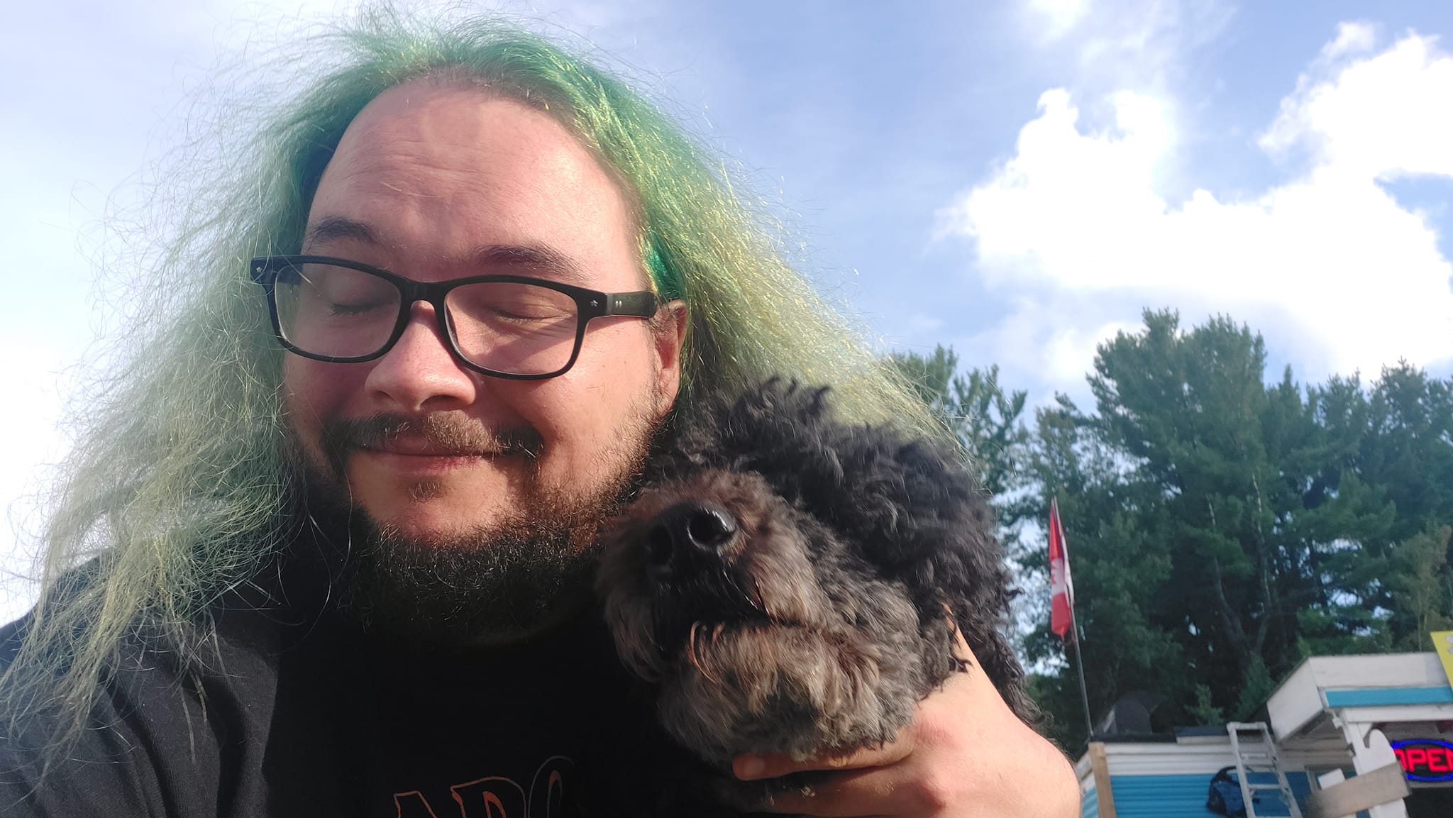 A man with green hair and a beard is hugging a black standard poodle who has booped him in her excitement